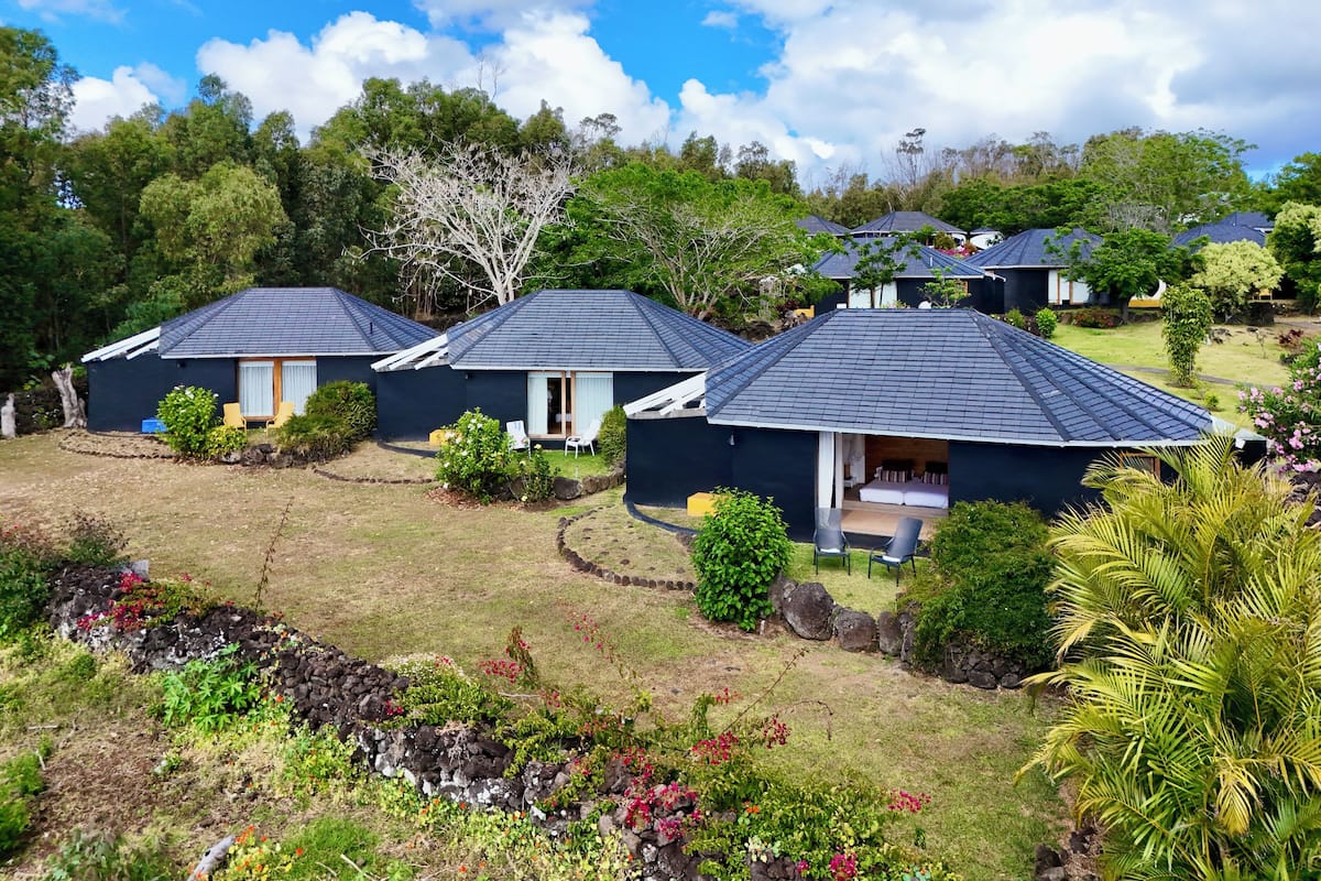 Fileira de cabanas azul-escuras com telhados inclinados, cercada por vegetação densa e árvores.