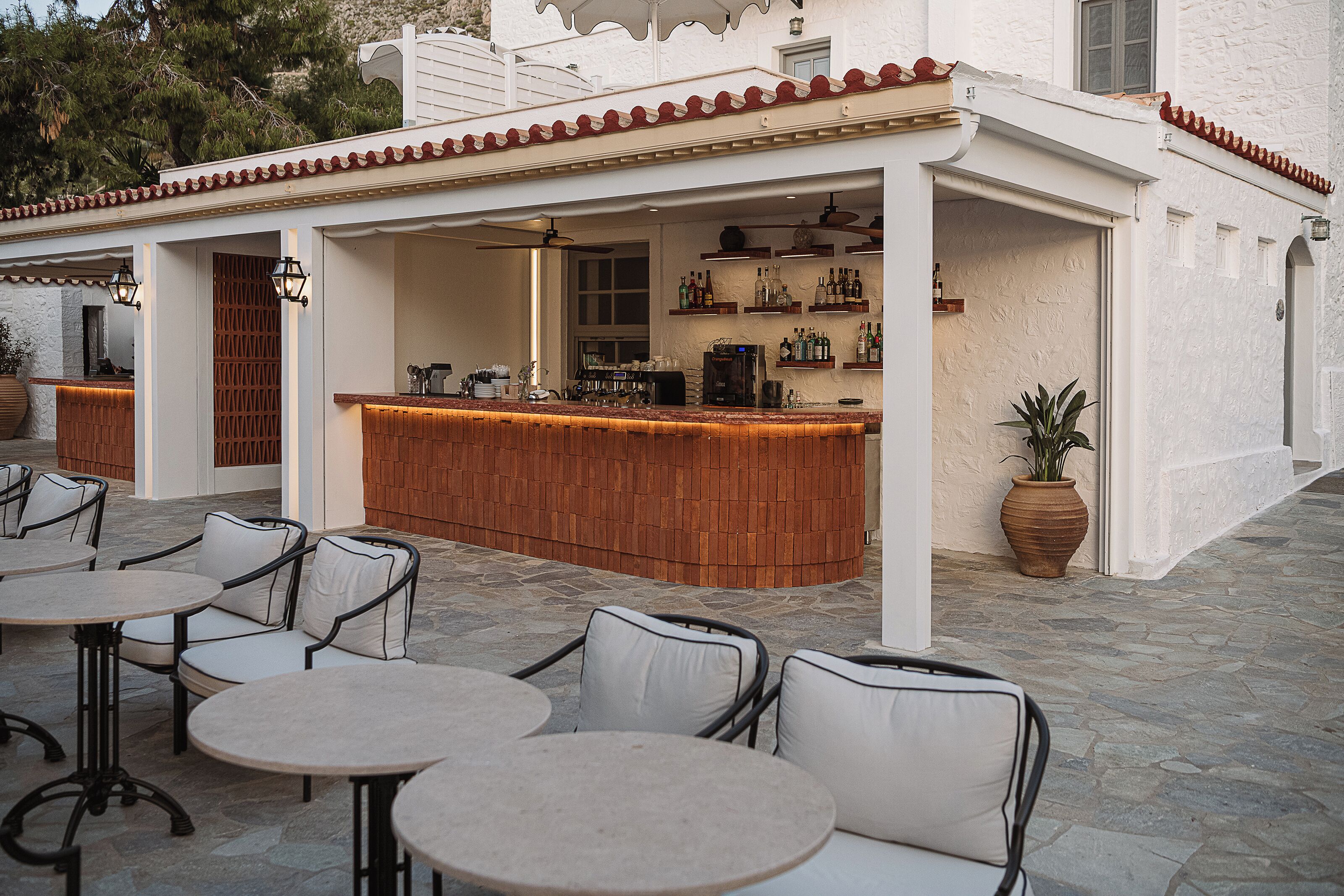 Una zona de bar con mostrador de madera, asientos exteriores con sillas blancas acolchadas y suelo de piedra.