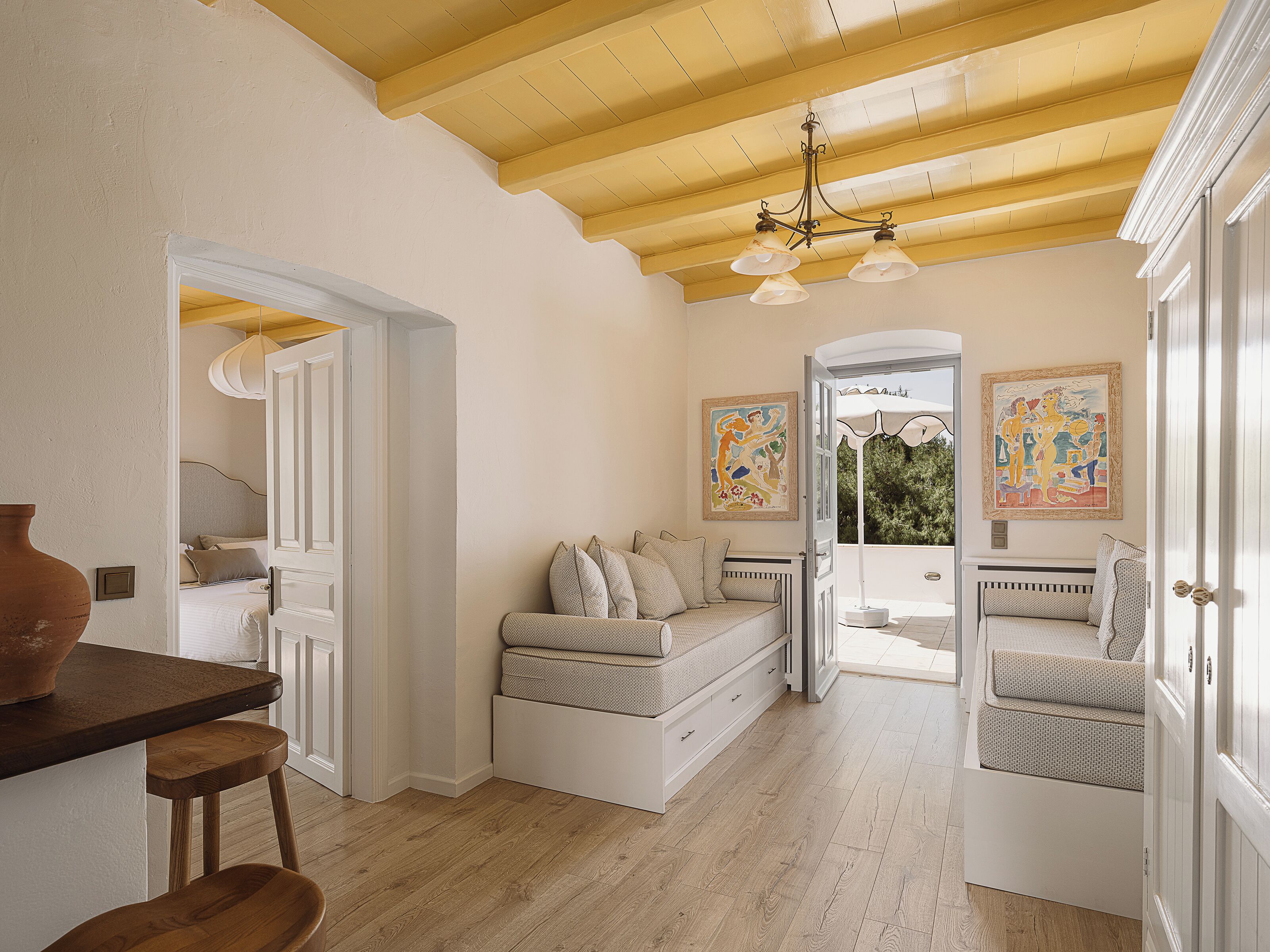 Una habitación con techo de madera, una cama con un banco, una mesa con sillas y obras de arte en las paredes.