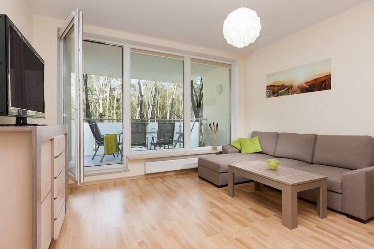 A modern living room with a sofa, a coffee table, a TV, and a sliding glass door leading to an outdoor area with chairs.