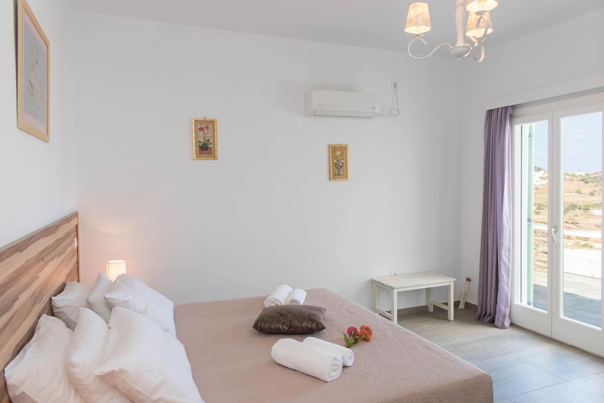 Un dormitorio con cama, almohadas, una lámpara de noche, una mesita y vistas a través de un ventanal.
