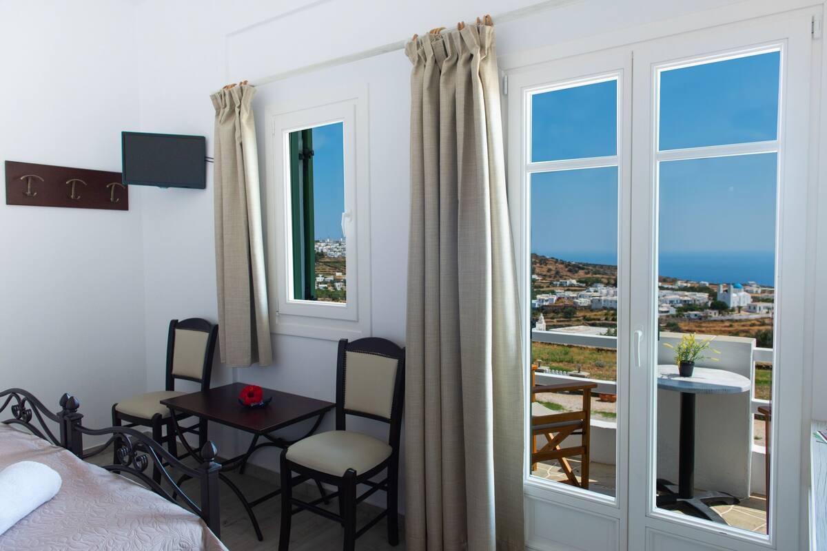 Habitación de hotel con cama, escritorio, dos sillas, una mesita con planta y vista al mar y edificios.