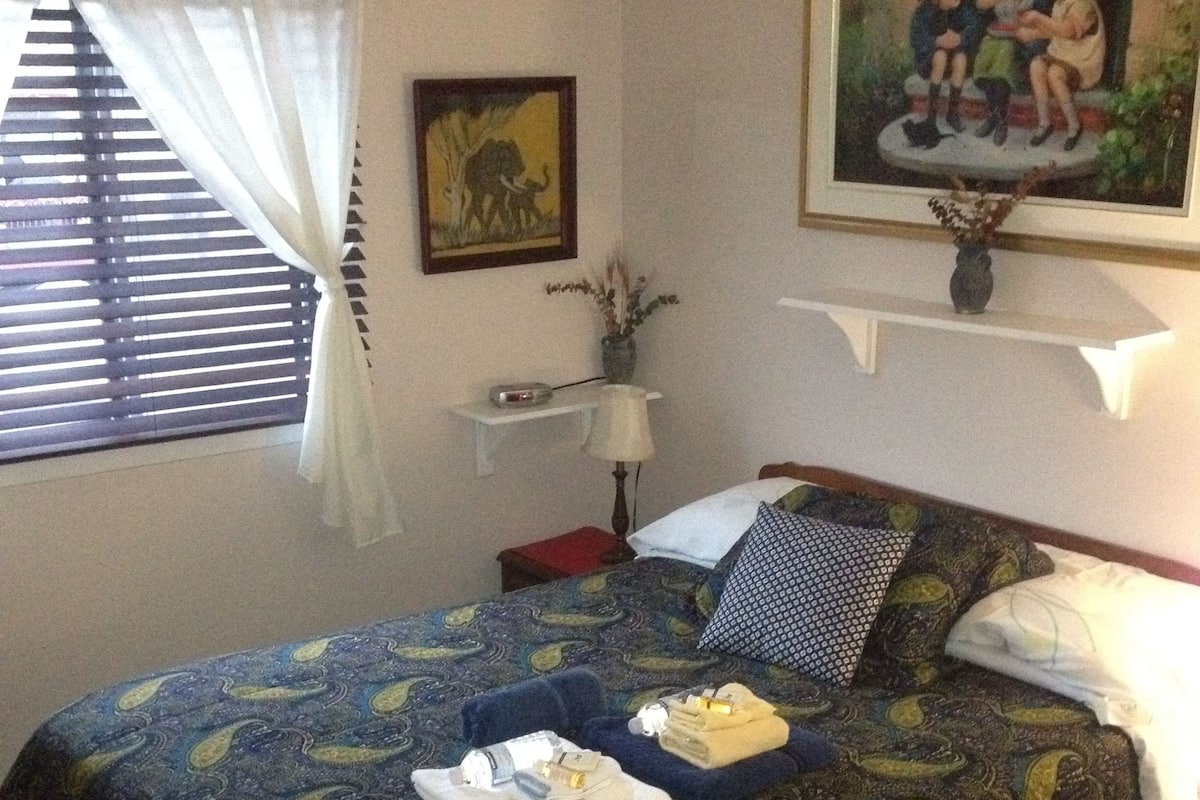 A bedroom with a bed, a nightstand, a lamp, a framed picture, and a window with blinds.