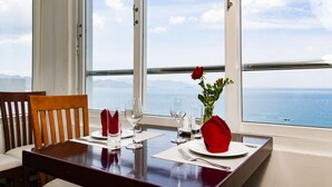 A dining table set with white plates, red napkins, and clear wine glasses, positioned near a window with a view of the sea. - Majestic Nha Trang Hotel (Nha Trang)