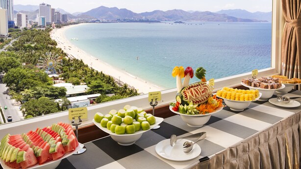 A buffet table with various fruits and desserts, a view of a beach and mountains, and a cityscape in the background. - Majestic Nha Trang Hotel (Nha Trang)