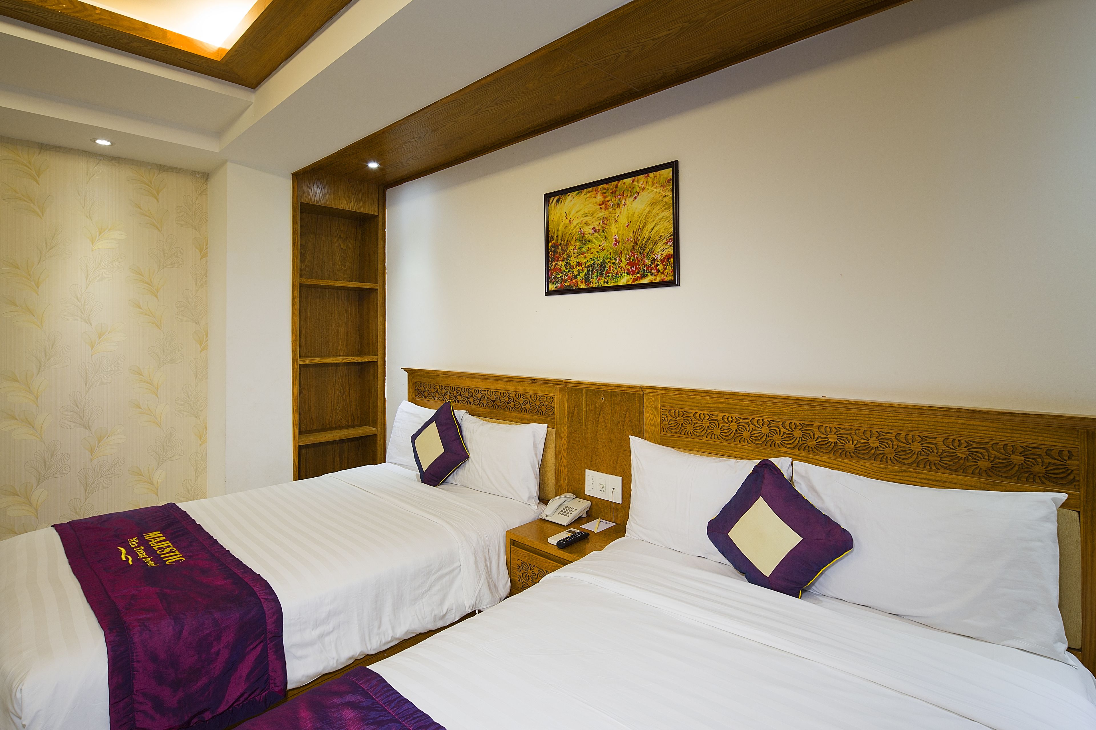 Une chambre d’hôtel avec deux lits, une tête de lit en bois, une table de chevet avec un téléphone et une télécommande, et un tableau décoratif au mur.