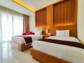 Quarto de hotel com duas camas, uma cadeira vermelha, uma mesa de cabeceira com luminária e uma janela com cortinas.