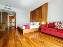 Una habitación de hotel con piso de madera, una cama con ropa blanca y un plaid rojo, un sofá rojo con cojines estampados, y una pared de paneles de madera.