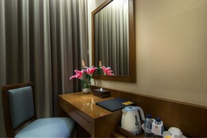 Quarto de hotel com escrivaninha de madeira, uma cadeira, um vaso com flores cor-de-rosa, um espelho, uma caixa de lenços, um caderno preto, uma chaleira e algumas garrafas de água.