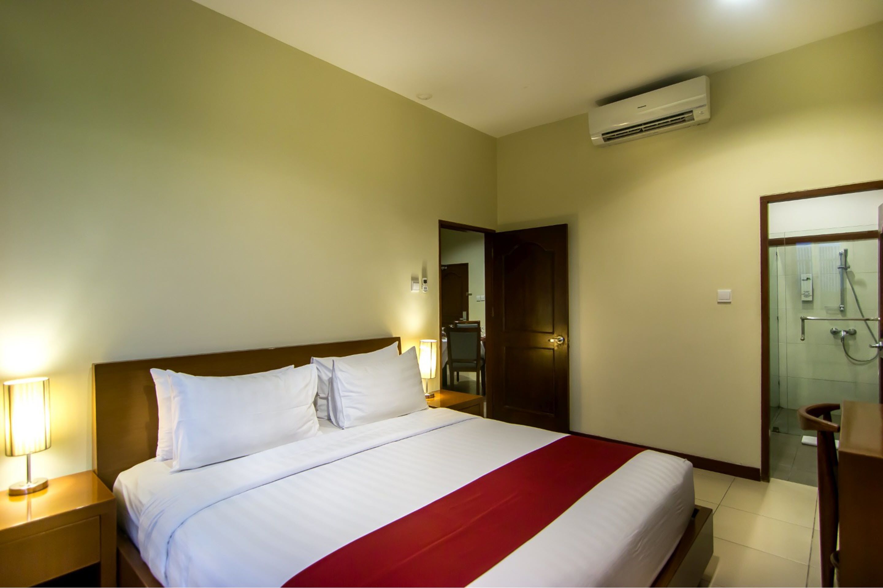 A hotel room with a bed, bedside lamps, a wooden headboard, a red bedspread, an air conditioning unit, a bathroom with a shower, and a wooden nightstand.