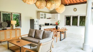 Una sala moderna con sofá, mesa de centro y comedor con sillas. Se ve una cocina al fondo con armarios y electrodomésticos blancos.