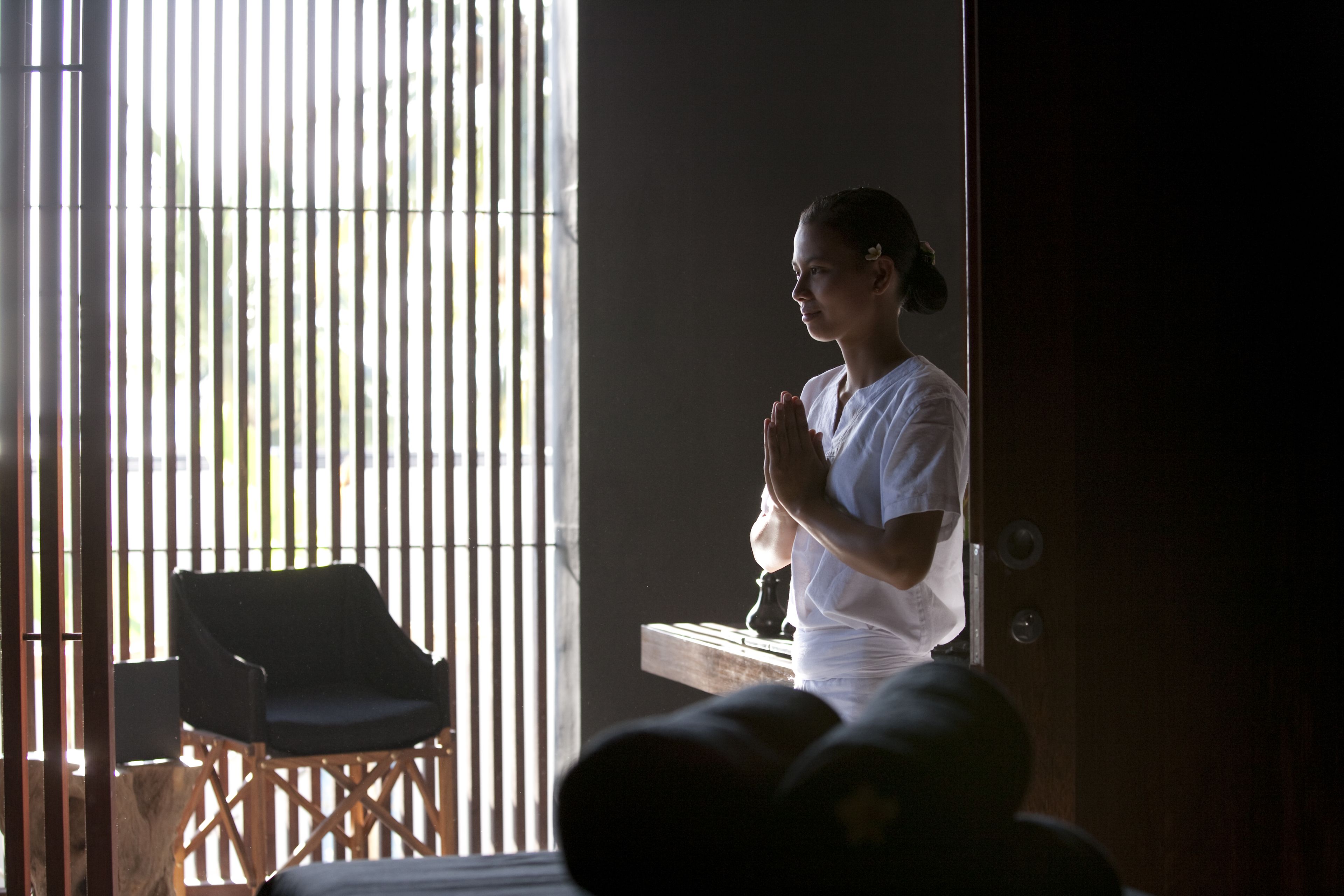 A person standing by a window with vertical blinds, a chair, and a small table with a decorative item.
