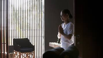 A person standing by a window with vertical blinds, a chair, and a small table with a decorative item.