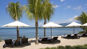 A beach with lounge chairs and umbrellas, palm trees, and a view of the ocean. - The Lombok Lodge Suites & Private Villas (Tanjung)