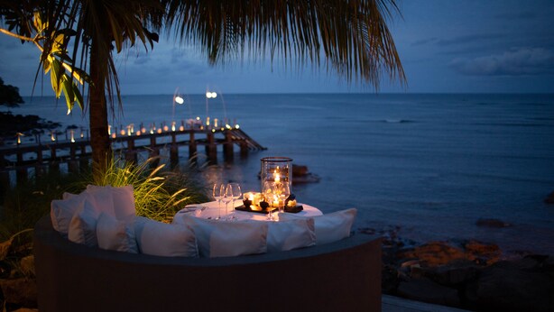 A table set with candles and glasses, overlooking a beach and a pier at dusk. - The Lombok Lodge Suites & Private Villas (Tanjung)