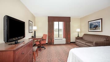A hotel room with a large flat-screen TV, a desk with a chair, a sofa, a bed, and a window with curtains.