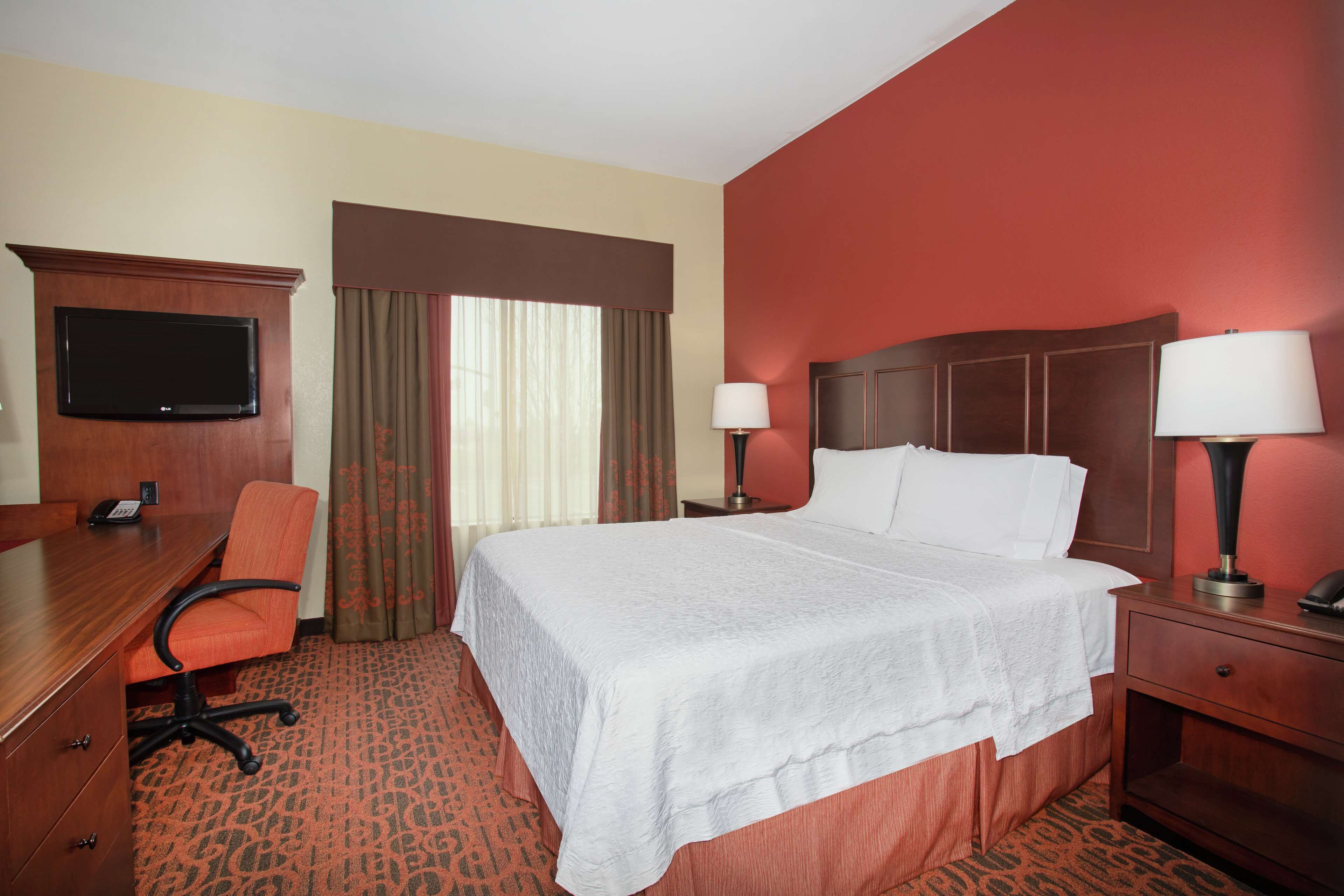 A hotel room with a large bed, a desk, a chair, a TV, two bedside lamps, and a window with curtains.
