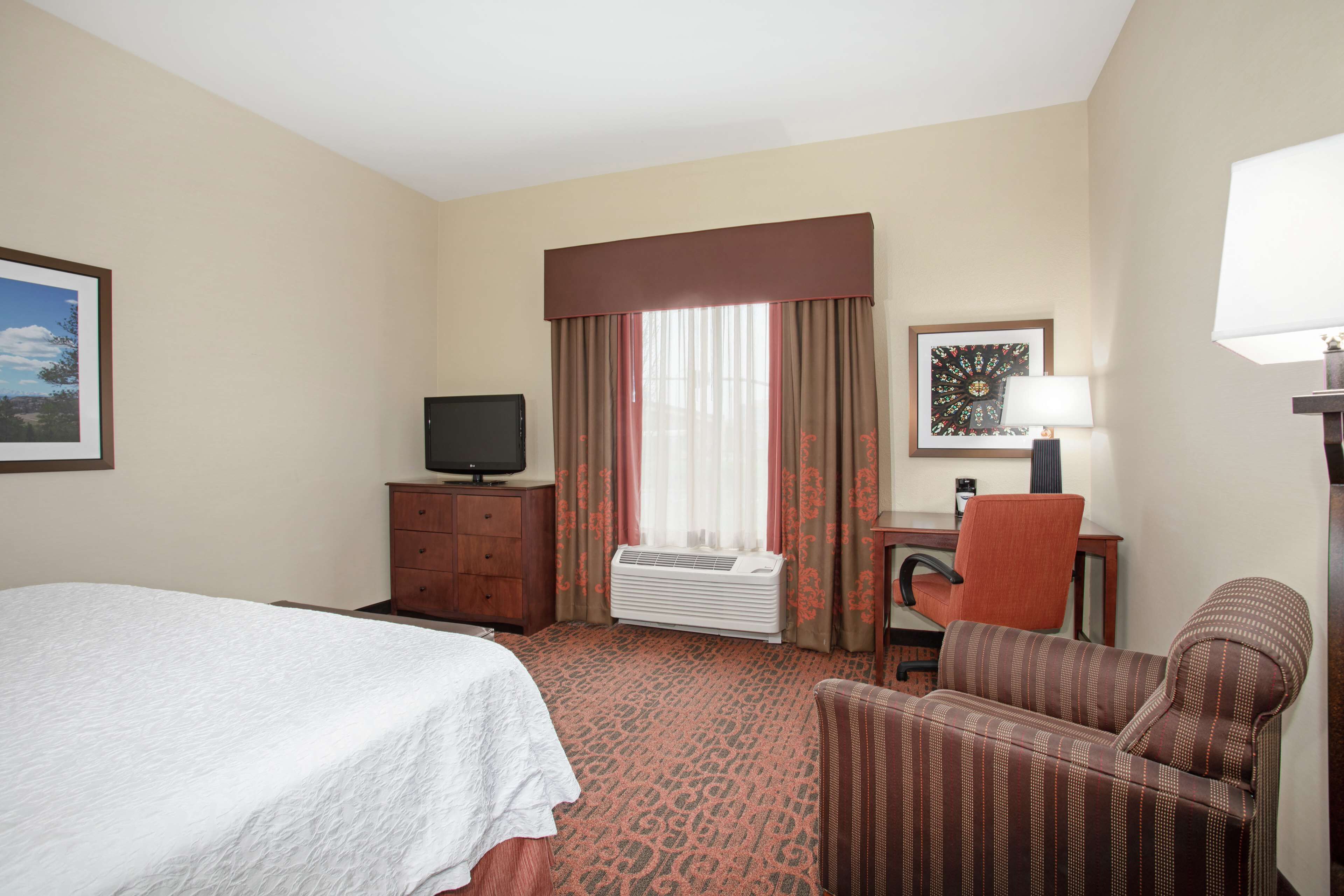A hotel room with a bed, a desk with a chair, a television, an air conditioner, and a window with curtains.