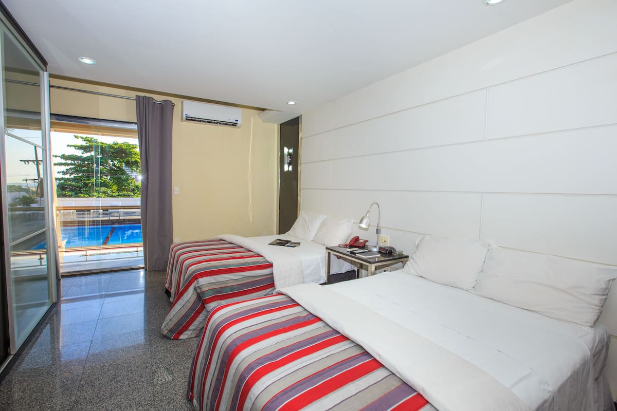 A hotel room with two beds, a bedside table with a lamp, a telephone, and a window overlooking a swimming pool.