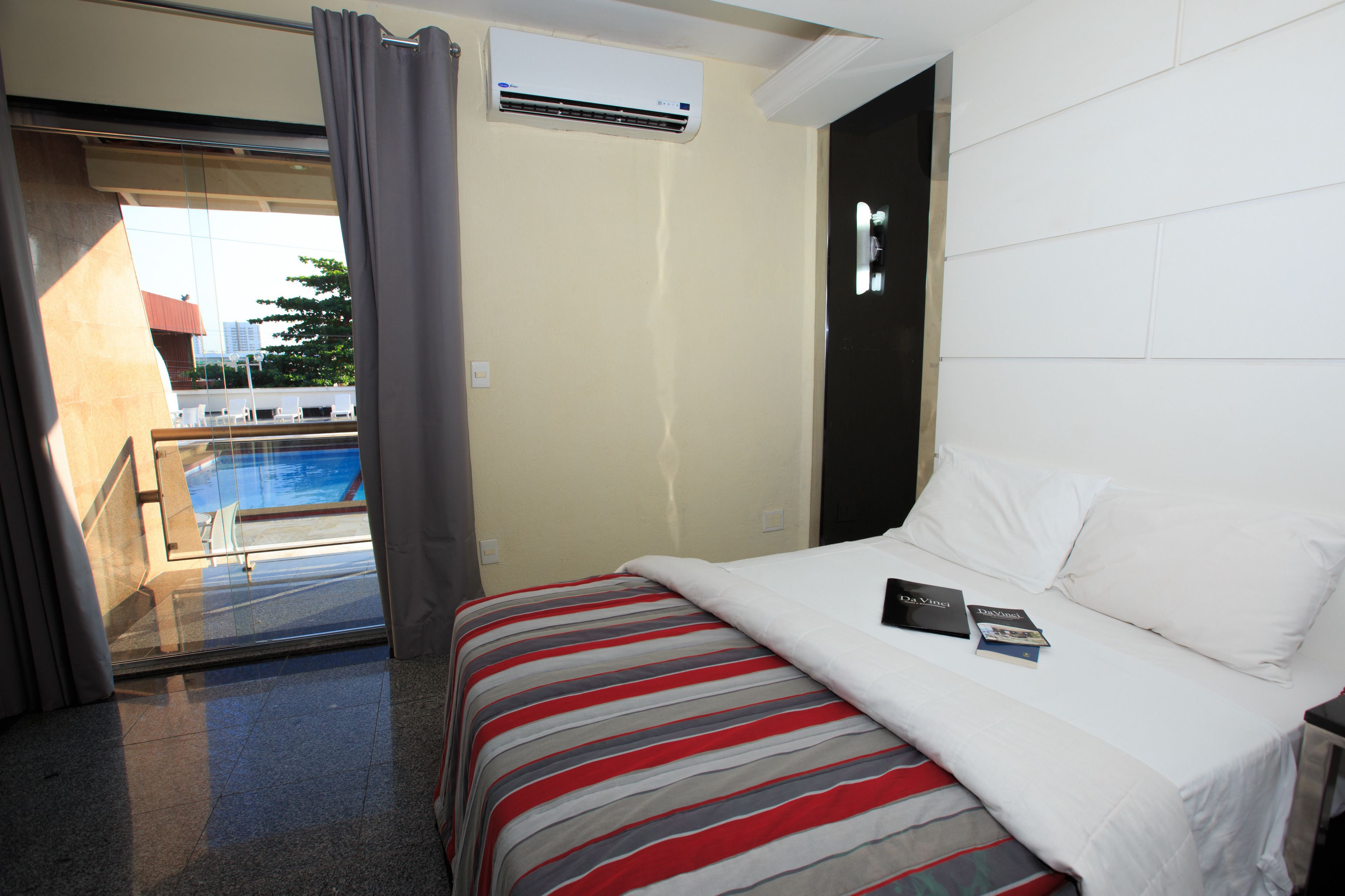 Habitación de hotel con cama, ventana con cortinas, equipo de aire acondicionado y vista a una piscina.