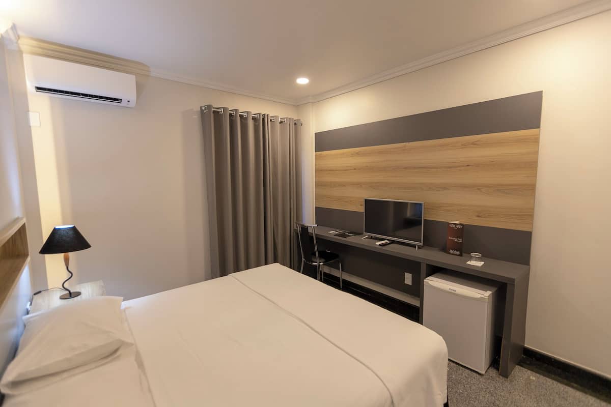 A hotel room with a bed, a desk with a computer, a TV, an air conditioner, a lamp, and a mini-fridge.