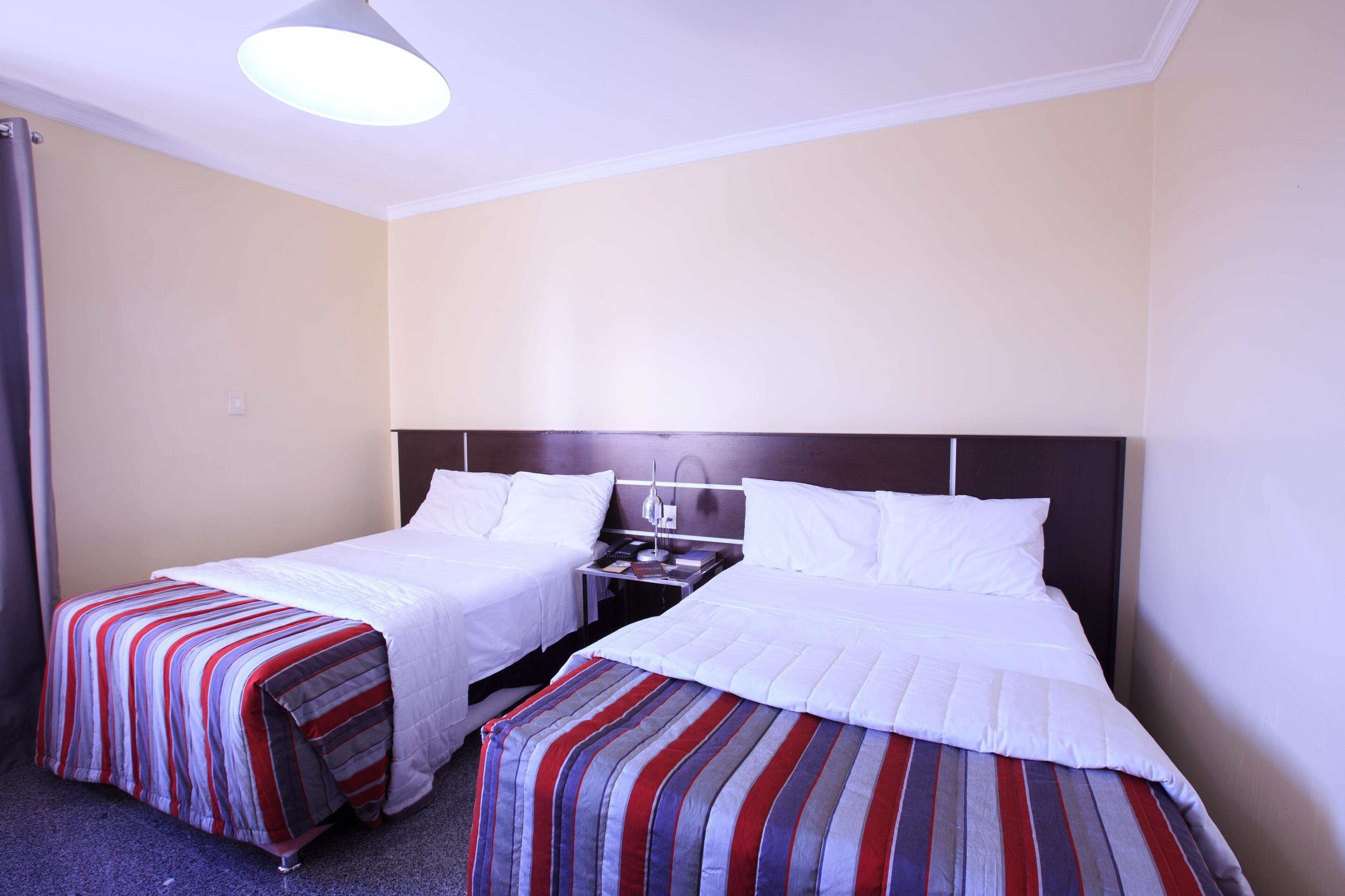 A hotel room with two beds, each with a striped bedspread, a bedside table with a lamp, and a ceiling light.