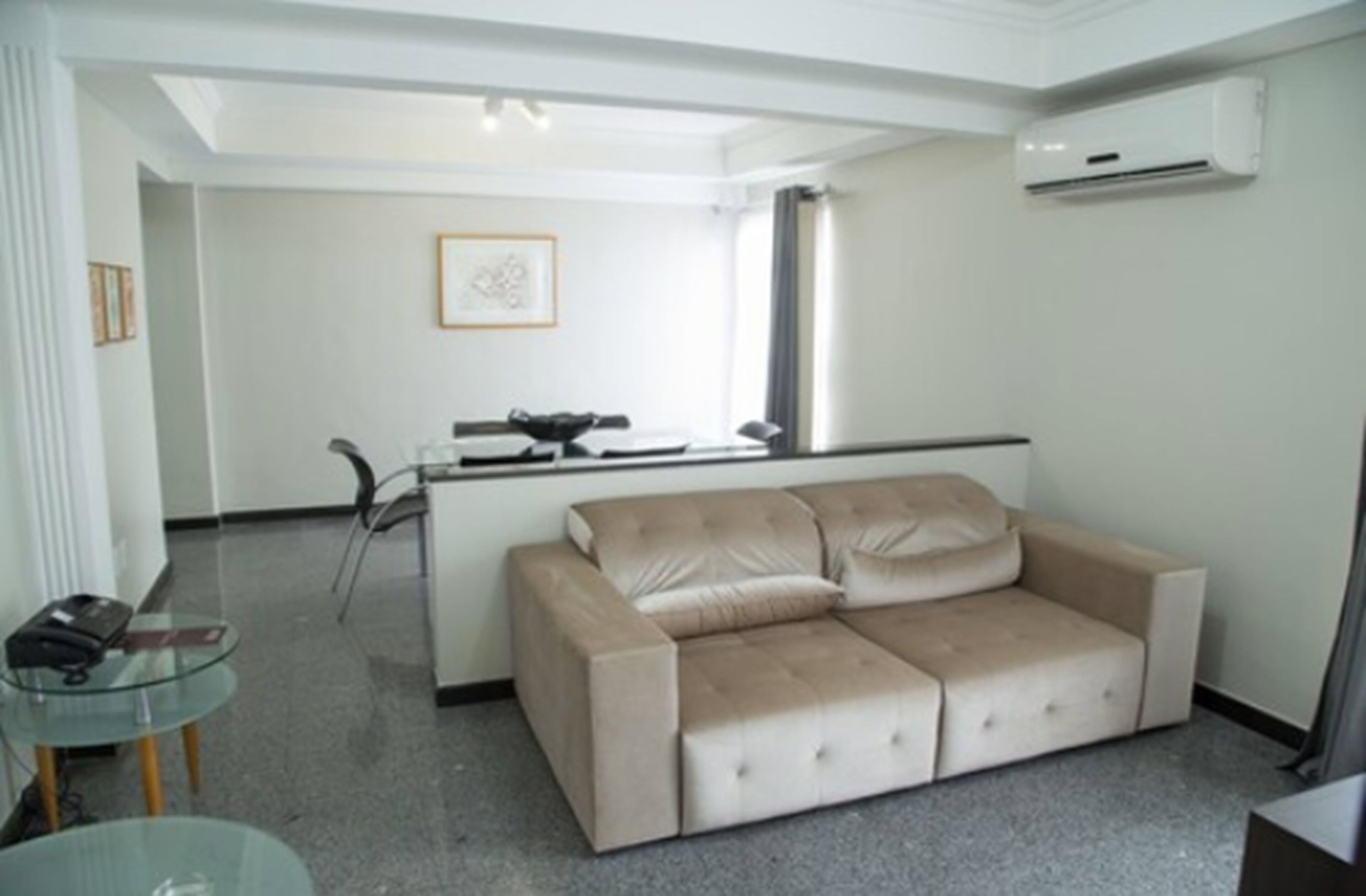A living room with a beige sofa, a glass coffee table, a small side table, and a kitchenette area with a microwave and a small refrigerator.