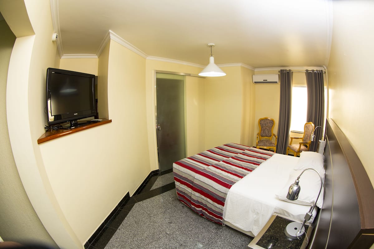 A hotel room with a bed, a television on a stand, a chair, and a desk with a lamp.