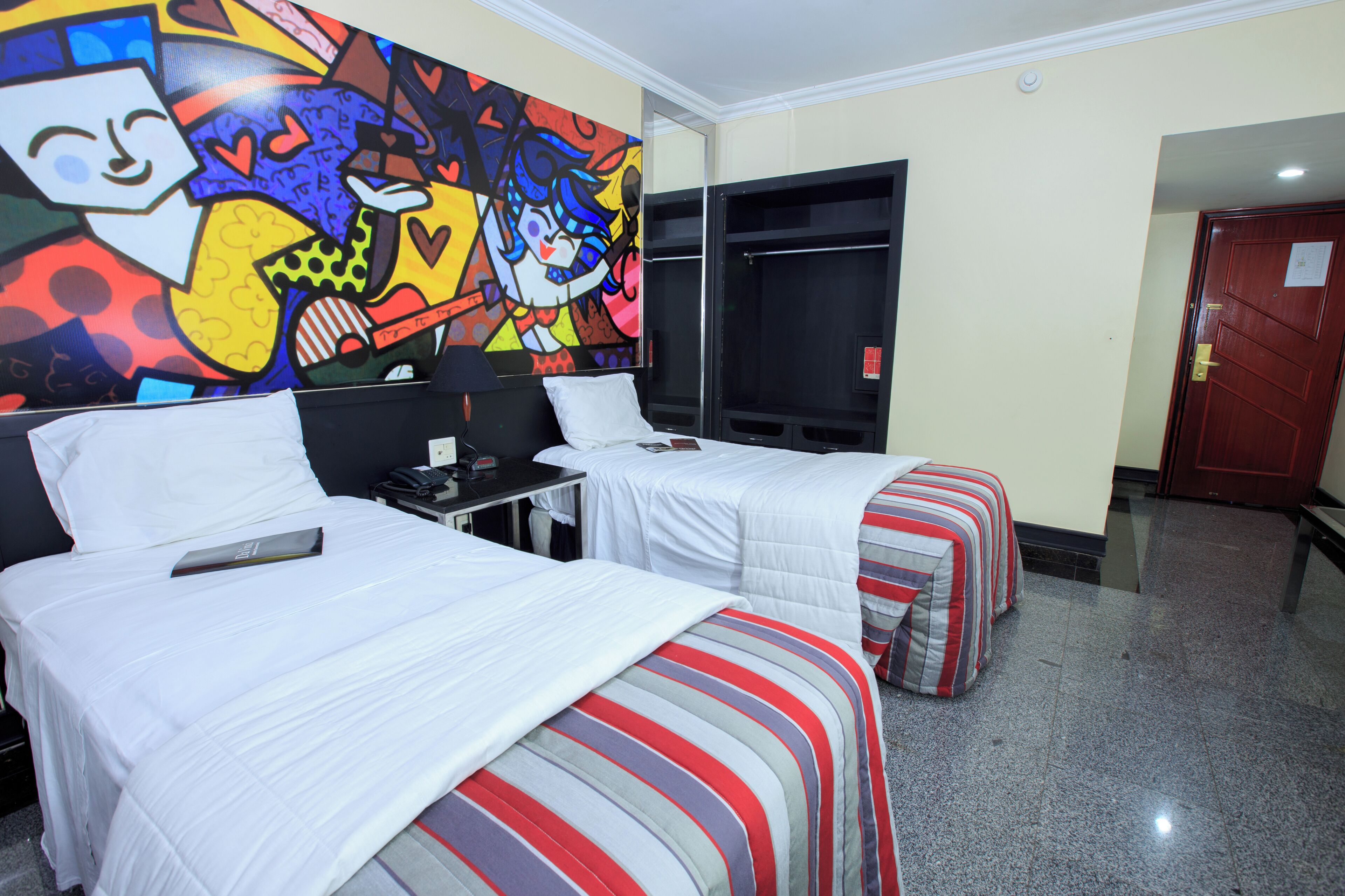 A hotel room with two beds, a large colorful mural on the wall, and a door to the right.