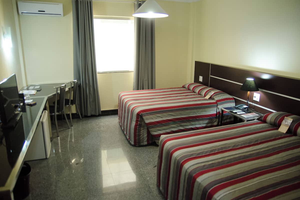A hotel room with two beds, a desk, a chair, a lamp, and a window with curtains.