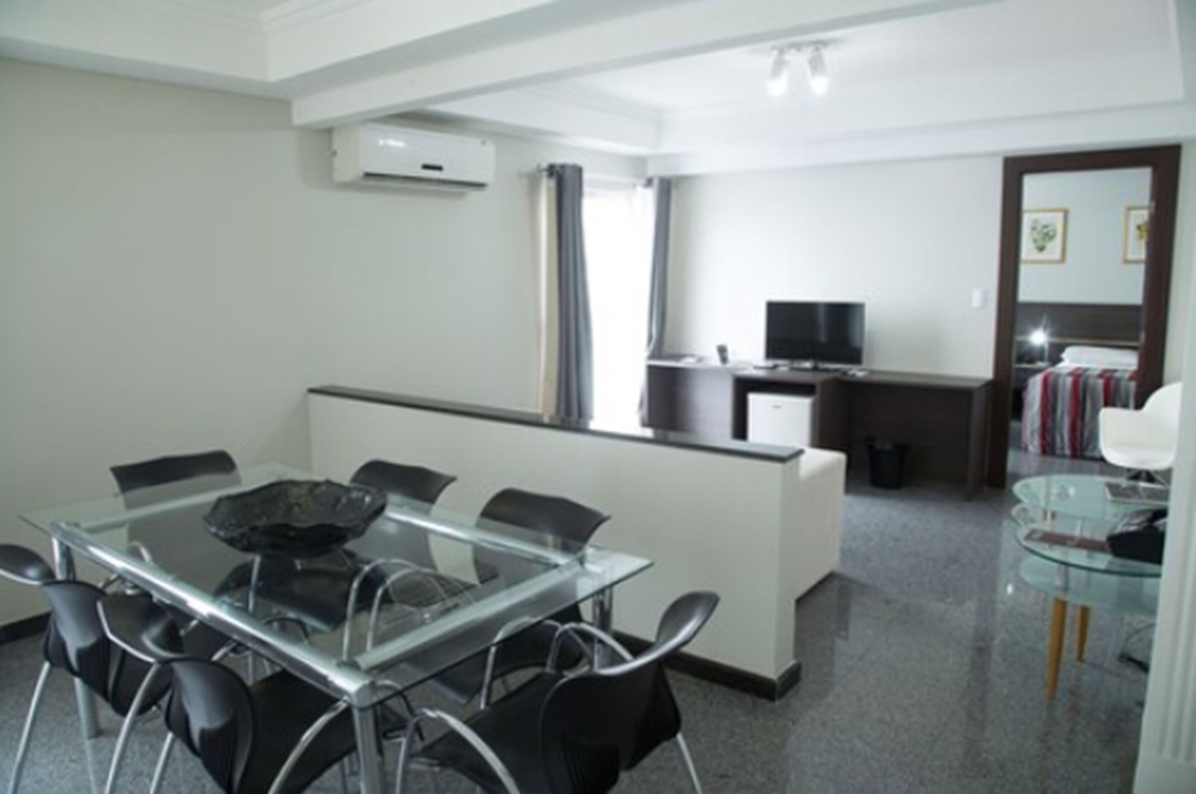 A modern hotel room with a glass dining table, black chairs, a small kitchen area, and a television.