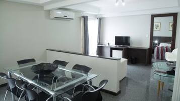 A modern hotel room with a glass dining table, black chairs, a small kitchen area, and a television.