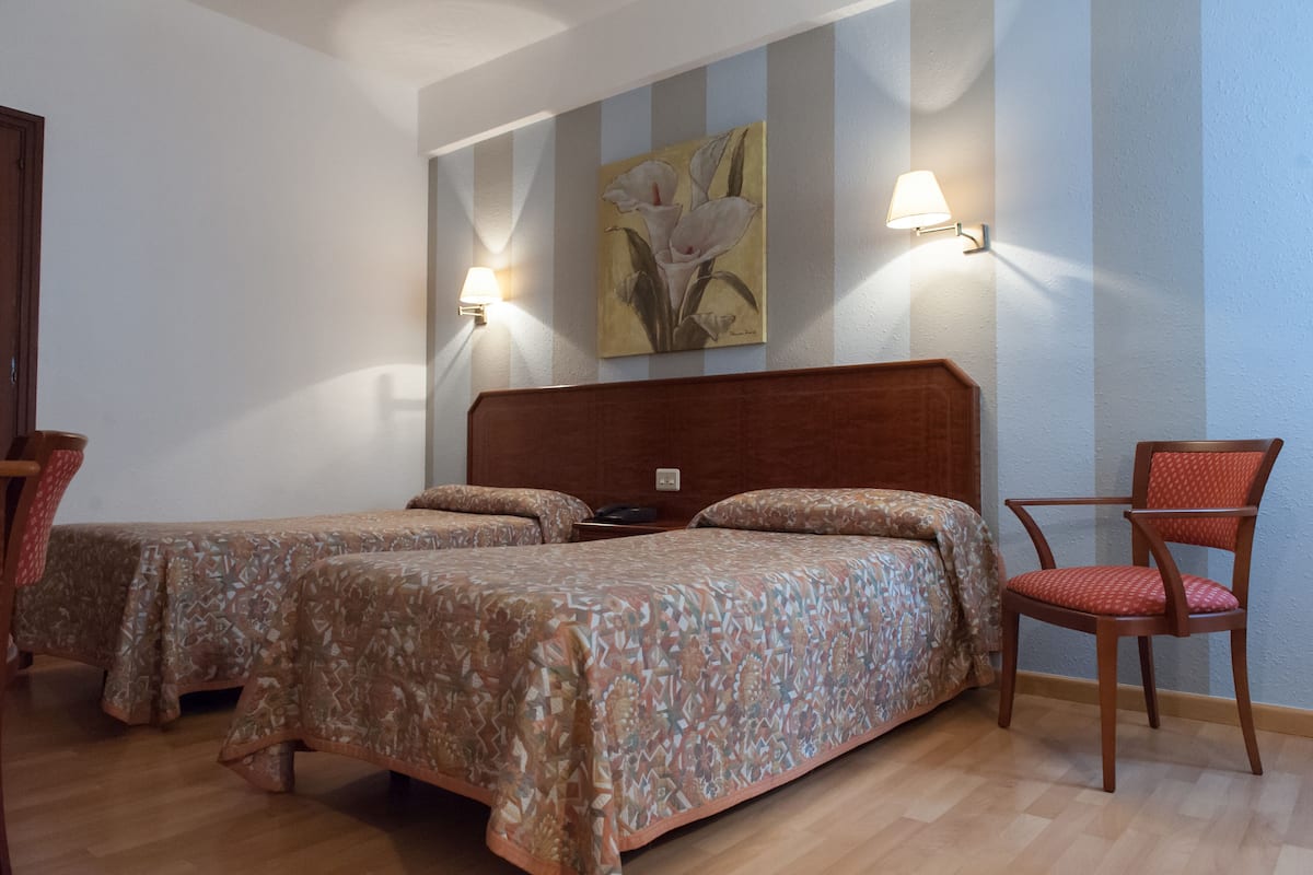 A hotel room with two beds, a wooden headboard, a chair, and a wall-mounted lamp.
