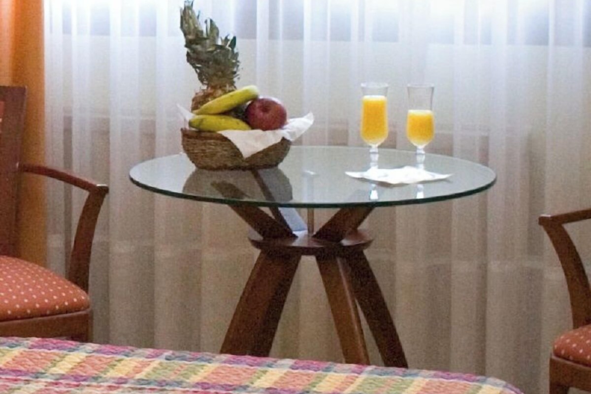 Habitación de hotel con una mesa de vidrio dispuesta con fruta y jugo, un colorido edredón acolchado y sillas de madera.