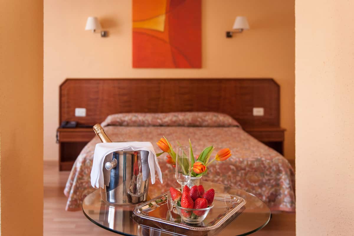 A hotel room with a bed, a bedside table with a champagne bottle, a tray with strawberries and tulips, and a wall with a red abstract painting.