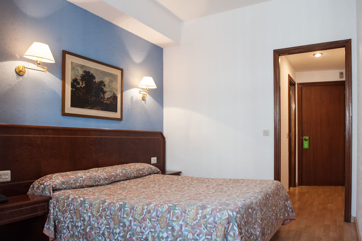 A hotel room with a wooden headboard, two bedside lamps, a framed picture on the wall, and a door leading to another room.