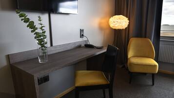 Ett hotellrum med ett skrivbord i trä, en gul stol, en krukväxt, en lampa och en tv som är monterad på väggen.