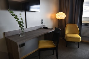 Habitación de hotel con un escritorio de madera, una silla amarilla, una planta en maceta, una lámpara y un televisor montado en la pared.