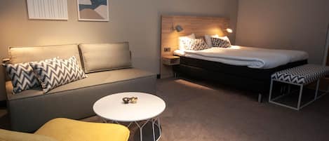 A modern hotel room with a sofa, a round coffee table, a bed with a headboard, and two wall-mounted artworks.