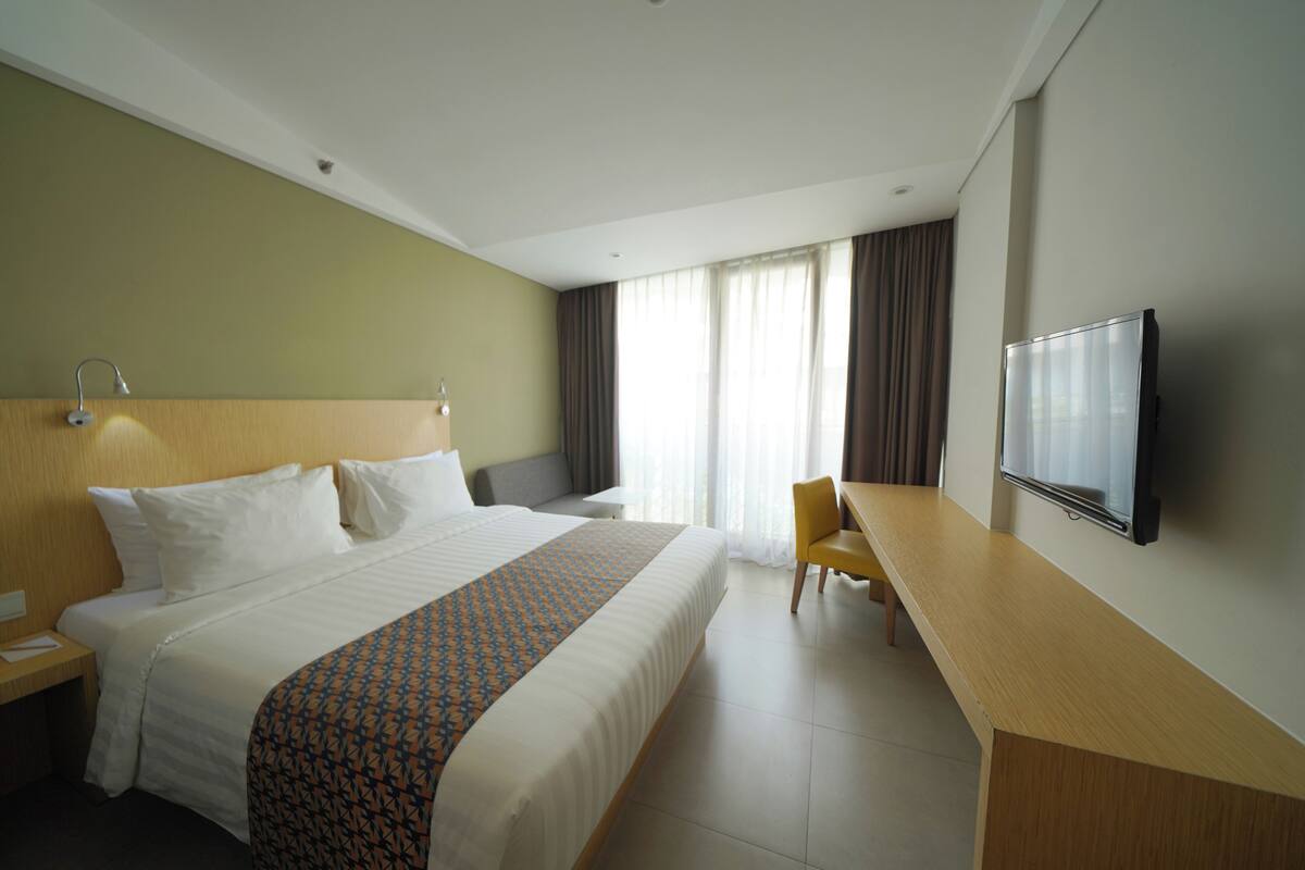 Une chambre d’hôtel avec un grand lit, un bureau, une chaise, une télévision et une fenêtre avec des rideaux.