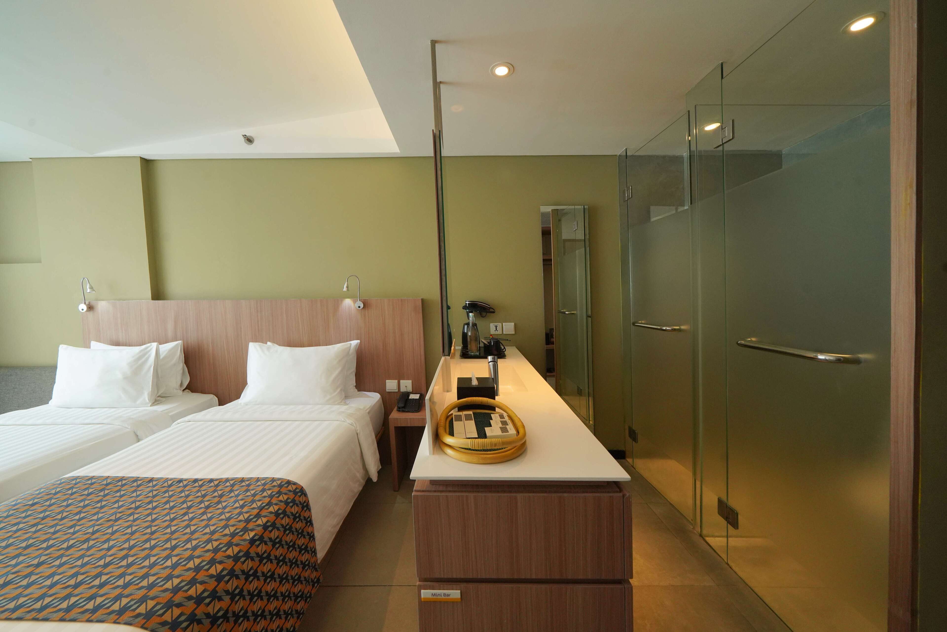A hotel room with two beds, a desk with a lamp, a minibar, and a glass-enclosed closet.