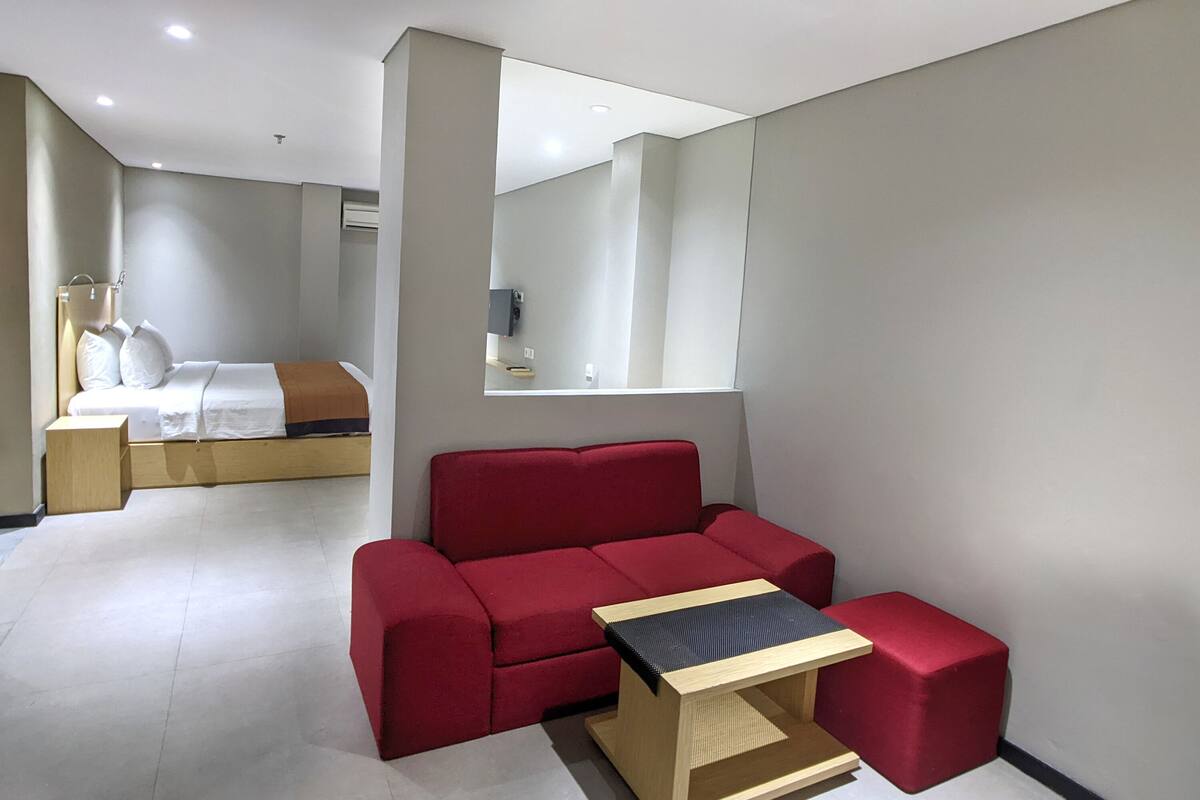 Une chambre d’hôtel moderne avec un canapé rouge, une table d’appoint en bois, un lit avec une literie blanche et un grand miroir au mur.