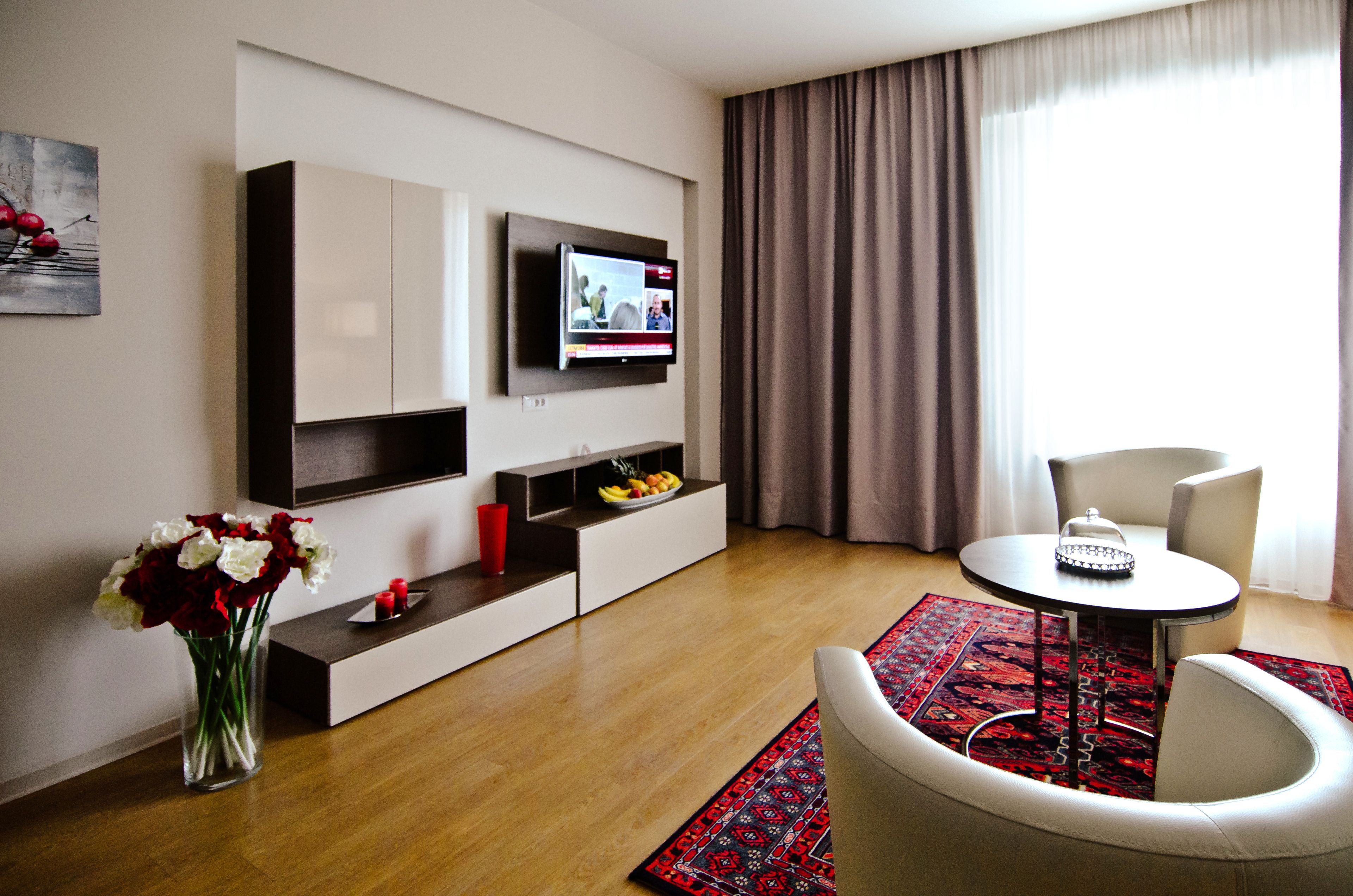 A modern living room with a flat-screen TV, a dining area with a table and chairs, a vase with flowers, and a fruit bowl on a shelf.