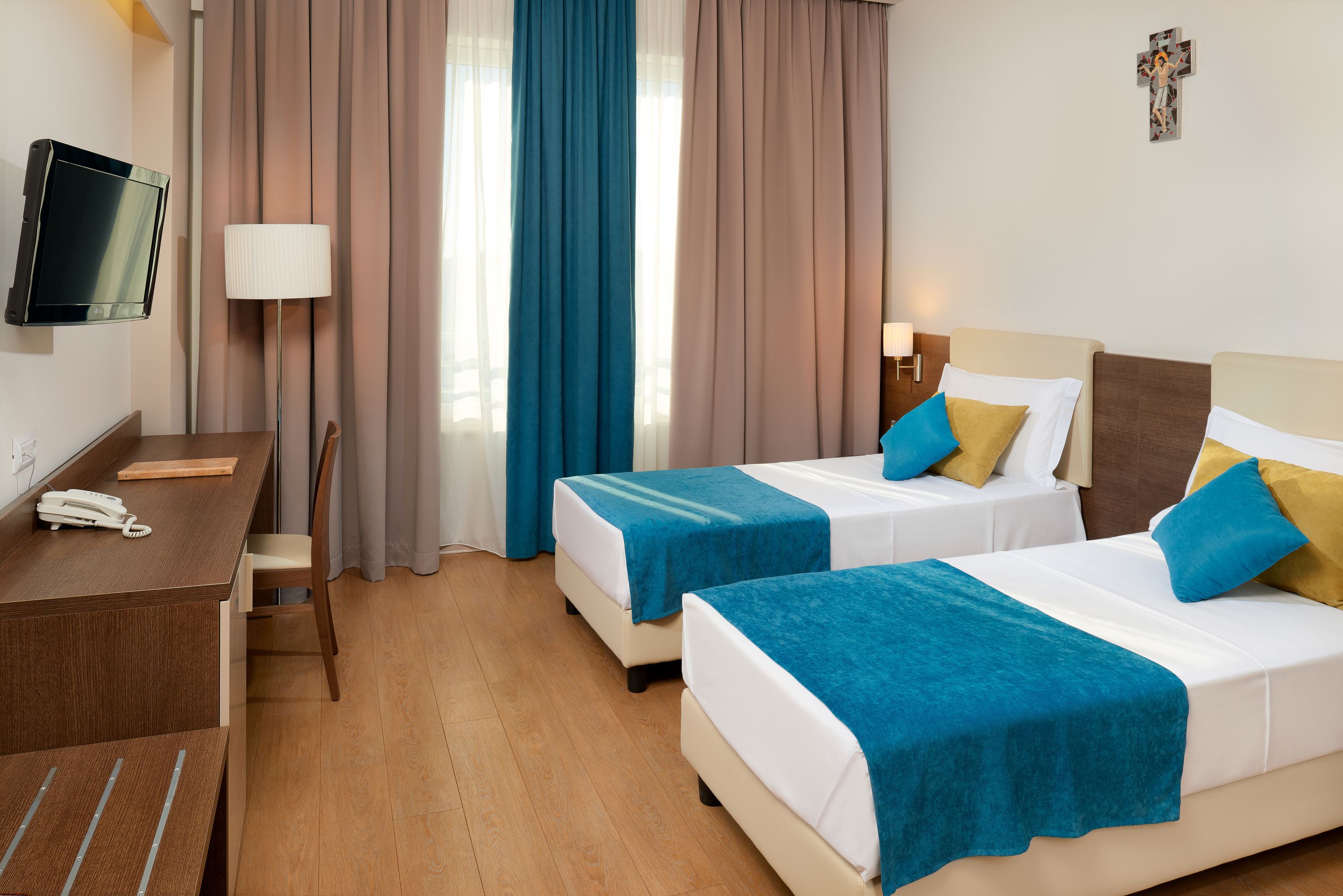 A hotel room with two beds, a desk with a phone, a TV, and a cross on the wall.