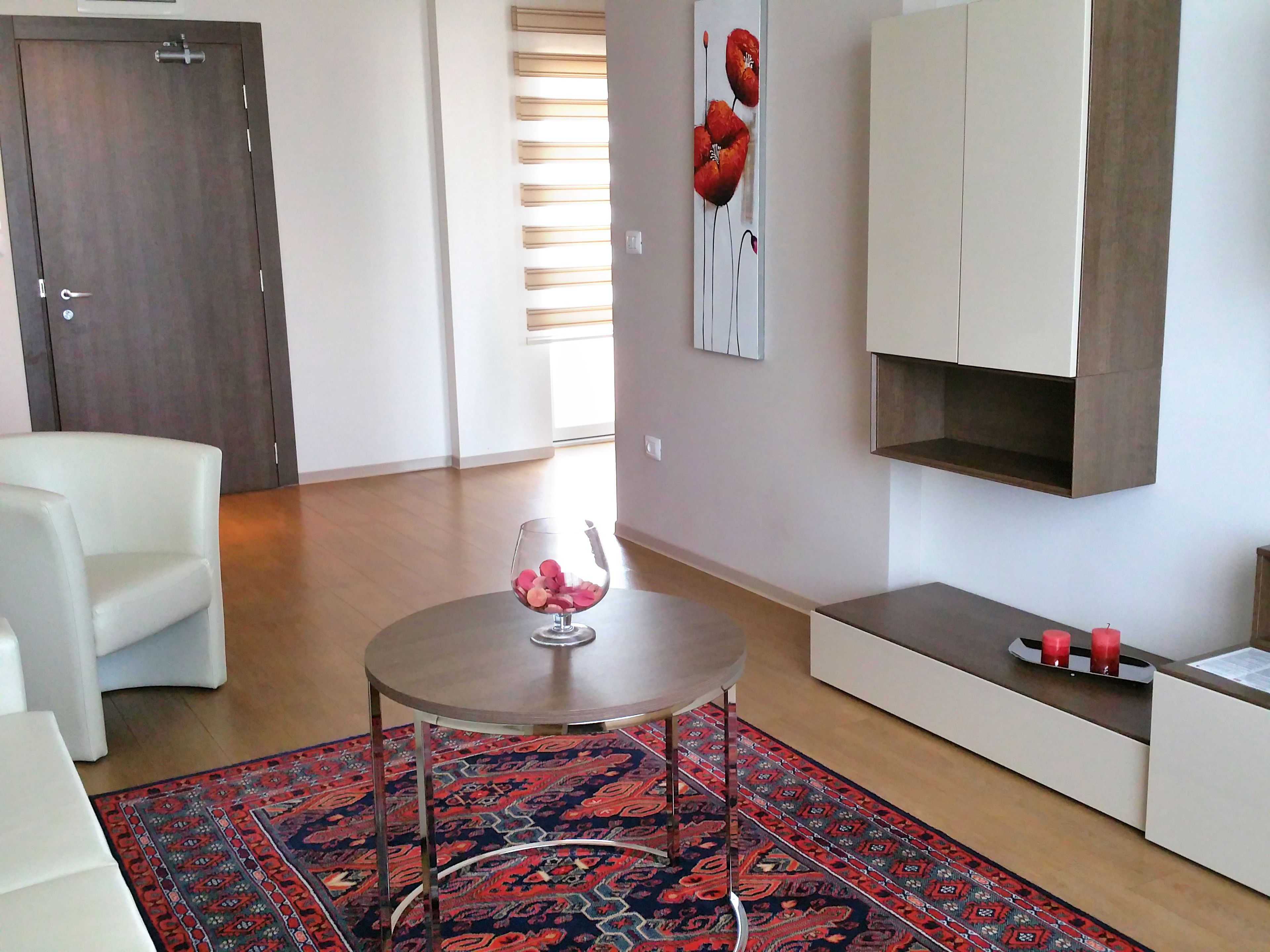 Un salon moderne avec un canapé blanc, une table ronde surmontée d’un vase en verre, un tapis à motifs et un meuble aux lignes épurées.