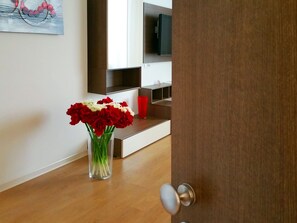 Una sala moderna con suelo de madera, un florero de vidrio con flores rojas y blancas, una taza roja y un televisor de pantalla plana sobre un estante de madera.