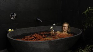 A woman in a large, dark-colored bathtub filled with water and rose petals, with a plant and a faucet visible in the background. - Jimbaran Cliffs Private Pool Hotel & Spa (Nusa Dua)