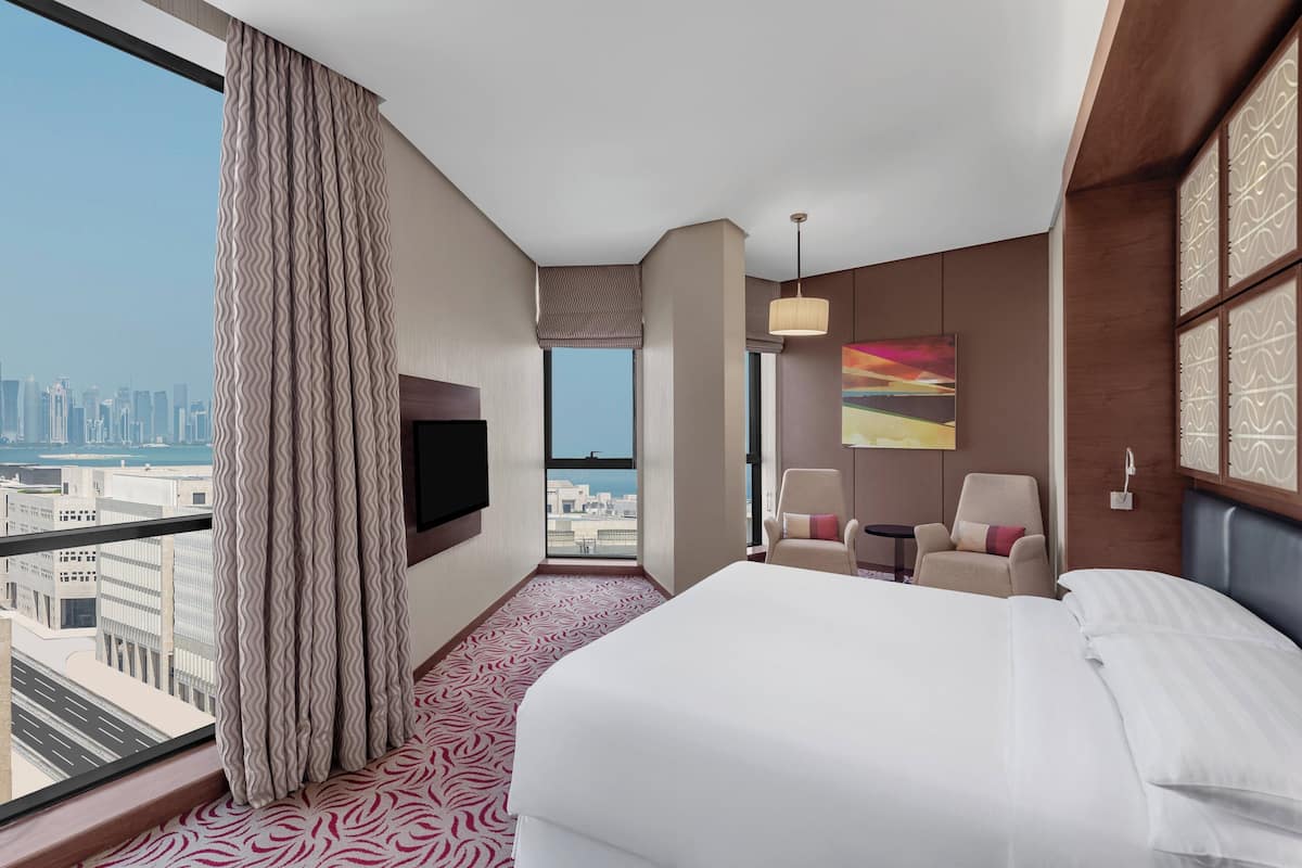 A hotel room with a large bed, a TV, a chair, and a view of the cityscape.