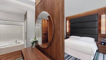 Una habitación de hotel moderna con tocador de madera, un espejo grande, una cama con ropa blanca y un baño separado con bañera.