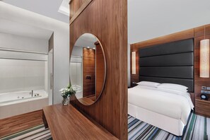 A modern hotel room with a wooden vanity, a large mirror, a bed with white linens, and a separate bathroom with a bathtub.